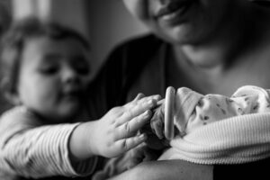 Die große Schwester streichelt dem Sternenkind liebevoll über seine Hände, das in den Armen der Mutter liegt.