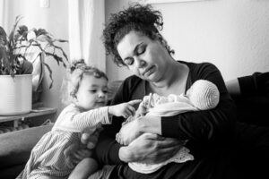 Eine Mutter sitzt auf dem Sofa und hält ihr Sternenkind, während das ältere Geschwisterkind sich an sie lehnt.