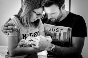 Ein Elternpaar steht eng beieinander und hält ihr in eine Decke gewickeltes Sternenkind liebevoll im Arm.