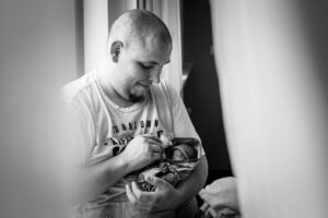 Ein Vater sitzt am Fenster und hält sein Sternenkind liebevoll in den Armen.