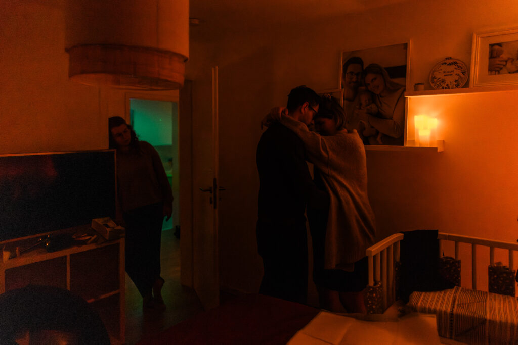 Gebärende wird nachts im Schlafzimmer von ihrem Partner gehalten während die Hebamme im Hintergrund begleitet